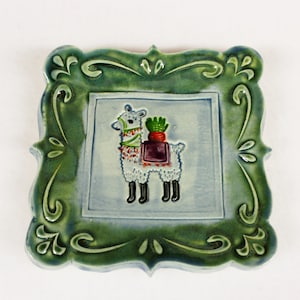 May include: Square ceramic tile with a green decorative border. The center features a light blue square with a white llama wearing a colorful harness and carrying a bundle of carrots. The llama has black feet.