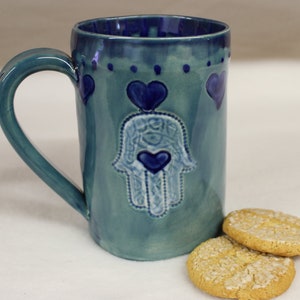 May include: A teal ceramic mug with a dark blue interior and handle. The mug features a hand-shaped design with a heart in the center and heart accents. Two golden cookies with a textured surface are next to the mug.