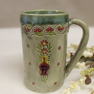 May include: A tall ceramic mug with a green rim and handle, featuring a floral design. The mug has a light green base with red polka dots and a central floral motif in red, yellow, and green. The rim has a decorative border.