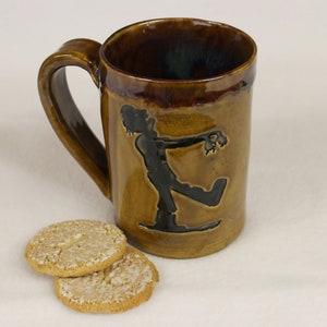 May include: A brown ceramic mug with a zombie silhouette design. The mug has a large handle and is accompanied by two round cookies. The mug is glazed with a glossy finish.