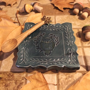 May include: A teal ceramic spoon rest with an owl design, a wooden spoon, and decorative wooden mushrooms and acorns. The spoon rest has a floral border. Autumn leaves surround the items, creating a fall theme.