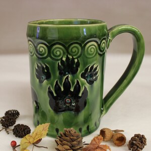 May include: A green ceramic mug with a large handle, decorated with black bear paw prints and a swirl pattern near the rim. The mug is approximately 5 inches tall and has a glossy finish.