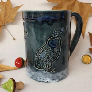 May include: A teal ceramic mug with a handle, featuring an owl design with stars and geometric patterns. The mug has a glossy finish and a white, foamy base. Decorative mushrooms and leaves surround the mug.