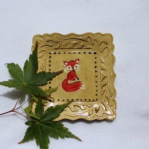 May include: A square, ceramic decorative plate with a raised, ornate border. The center features a red fox illustration on a tan background. The plate is tan with a glossy finish. Green leaves are in the foreground.