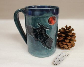 May include: A ceramic mug with a dark teal glaze, featuring a raised relief of a moth and a red circle. The mug has a large handle and a speckled pattern. A pine cone and a feather are next to the mug.