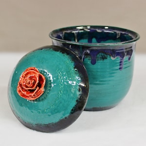 May include: A handcrafted ceramic jar with a lid. The jar is teal with dark blue accents and a red rose on the lid. The lid is slightly ajar, revealing the interior. The jar has a glossy finish.
