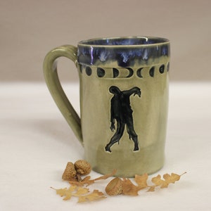 May include: A ceramic mug with a green handle and body, featuring a black zombie silhouette. The rim is dark blue with a moon phase design. Acorns and leaves are scattered near the base of the mug.