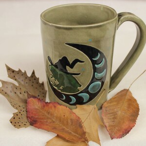 May include: A sage green ceramic mug with a black crescent moon design featuring a witch silhouette and teal accents. The mug has a curved handle and is decorated with small teal dots. Dried leaves are arranged around the mug.