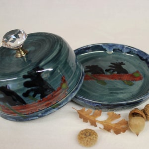 May include: A handcrafted ceramic butter dish and plate set. The teal-colored dish features a canoe scene with black figures and a red canoe. The lid has a crystal knob. The plate also has the canoe scene. Decorative fall leaves and acorns are next to the dish.