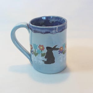 May include: A light blue ceramic mug with a handle. The mug features a black silhouette of a rabbit, flowers, and a white fence design. The rim and handle are a darker shade of blue.