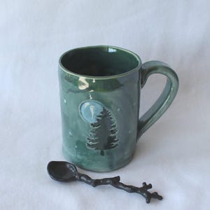 May include: A handmade green ceramic mug with a tree and moon design, accompanied by a dark spoon. The mug features a handle and is decorated with white dots. The spoon has a twig-like handle.