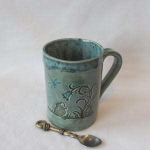 May include: A cylindrical ceramic mug with a glossy sage green glaze and a darker blue-green rim. The exterior features a raised relief design of a small bird, a flower, and a dragonfly. A small, twisted-handle metal spoon rests in front.