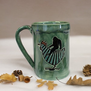 May include: A green ceramic mug with a large handle. The mug features a black silhouette of Bigfoot riding a chicken. The chicken has blue and black striped feathers. The mug is decorated with a glossy glaze.