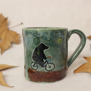May include: A ceramic mug with a green and brown glaze. The mug features a black bear riding a bicycle with a basket of flowers, and a yellow sun. The mug is surrounded by brown autumn leaves.