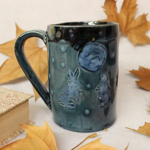 May include: A ceramic mug with a teal glaze, featuring a dark blue rim and handle. The mug is decorated with embossed blue rabbits, a moon, and small dots. The mug is approximately 5 inches tall.