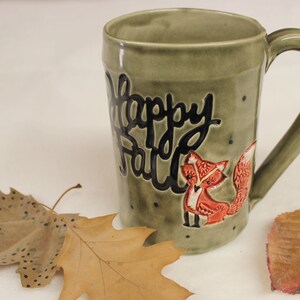 May include: A ceramic mug with a green glaze featuring the words "Happy Fall" in black script and a red fox illustration. The mug has a large handle and is decorated with small black dots. Dried leaves are in the foreground.