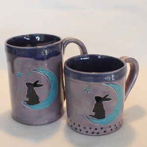 May include: Two ceramic mugs with a whimsical design. The mugs are lavender with a dark blue rim and handle. Each mug features a black rabbit silhouette sitting on a turquoise crescent moon, with stars and dots.