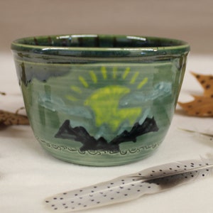 May include: A green ceramic bowl with a hand-painted landscape design featuring a yellow sun, mountains, and clouds. The bowl is surrounded by autumn leaves, acorns, and feathers.