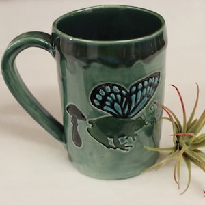 May include: Green ceramic mug with a butterfly and frog design. The mug has a large handle and a glossy finish. The butterfly has blue and black wings. A small plant with red-tipped leaves is next to the mug.