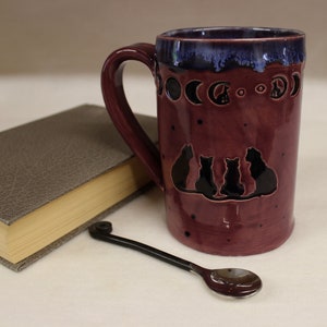 May include: A burgundy ceramic mug with a dark blue rim and handle. The mug features a design of black cat silhouettes and moon phases. A small black spoon rests beside the mug and a closed book.