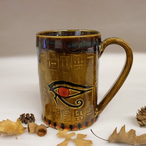 May include: A brown ceramic mug with a handle, featuring an Eye of Horus design and hieroglyphs. The mug has a glossy finish with a dark brown rim and a red, black, and gold color scheme. The mug is approximately 5 inches tall.