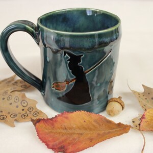May include: A teal ceramic mug with a black witch silhouette riding a broom. The mug has a curved handle and is decorated with autumn leaves and an acorn. The mug is approximately 4 inches tall.