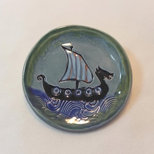 May include: A round, ceramic dish with a Viking ship design. The ship is black with a striped sail, set against a blue and green background. The rim of the dish is a darker green.
