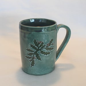 May include: A teal ceramic mug with a handle. The mug features a hand-painted design of a holly branch with green leaves and red berries. The mug has a glossy finish and is suitable for hot or cold beverages.