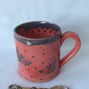 May include: A coral-colored ceramic mug featuring a dark purple rim with a drip pattern, embossed bat motifs, and small black dots. The mug has a sturdy handle and is accompanied by a small, metallic, twisted-handle spoon with a loop end.