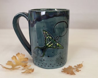 May include: A teal ceramic mug with a large handle. The mug features a green and yellow moth design with black details. The mug has a glossy finish and is decorated with small black dots and swirls.