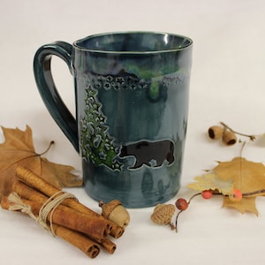 May include: A teal ceramic mug with a bear and Christmas tree design. The mug has a large handle and a glossy finish. The mug is decorated with a black bear silhouette and a green Christmas tree with star accents.