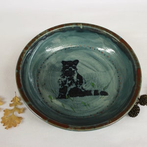 May include: Handmade ceramic bowl with a teal interior and a raccoon illustration. The bowl has a brown rim and is decorated with small red dots. The raccoon is black and white. The bowl is approximately 8 inches in diameter.