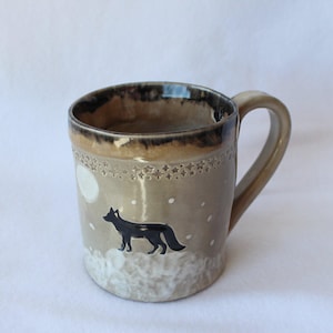 May include: A ceramic mug with a glossy beige finish featuring a dark brown rim. The exterior displays a black fox silhouette standing on a white textured base, a white circular moon, and a band of small star-shaped indentations near the top.
