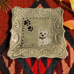 May include: A square, sage green ceramic dish with a decorative border. The center features a cartoon dog and paw prints. The dish has a raised design and is set against a patterned background.