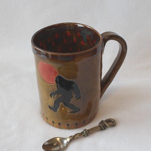 May include: A brown ceramic mug with a glossy finish featuring a black silhouette of a creature walking toward a red circular sun. The interior and base are decorated with small red teardrop shapes. A small, twisted metal spoon lies in front.