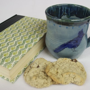 May include: A ceramic mug with a blue bird design, a book with a green and white patterned cover, and two cookies. The mug has a blue glaze and a handle. The book is closed, and the cookies are light brown.