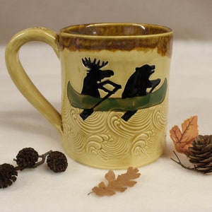 May include: A ceramic mug with a light yellow base and a brown rim. The mug features a black silhouette of a moose and a bear in a green canoe, with a wave pattern. The handle is light yellow.