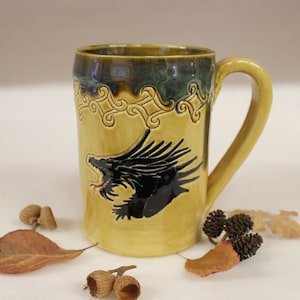 May include: A handmade ceramic mug with a dragon design. The mug is primarily yellow with a black dragon illustration and a decorative border. The handle and rim are a darker yellow and green. Acorns and leaves are scattered around the mug.