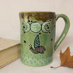 May include: A green ceramic mug with a handle, decorated with a snail with butterfly wings, crescent moons, and stars. The mug has a brown rim and a light green body. A book and a leaf are in the background.
