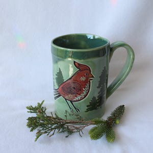May include: A green ceramic mug with a red bird design and forest silhouettes. The mug has a curved handle and a glossy finish. The bird is detailed with intricate patterns. A sprig of greenery rests near the mug.