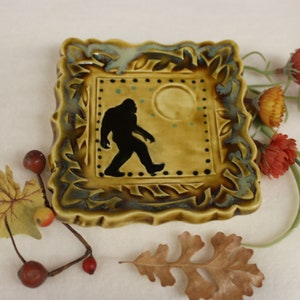 May include: A square, ceramic Bigfoot-themed decorative tile. The tile features a black silhouette of Bigfoot walking across a tan background with a yellow moon and teal dots. The tile is framed with a green and brown leaf design.