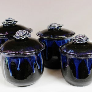 May include: Four ceramic jars with lids, each adorned with a rose-shaped handle. The jars are a deep, glossy black with vibrant blue drips, creating a striking contrast. These decorative containers are ideal for storing small items.