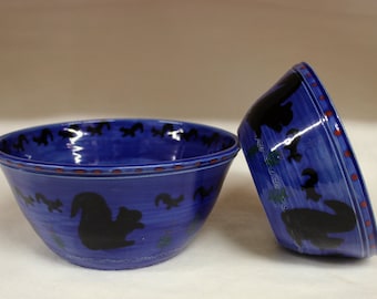 May include: Two blue ceramic bowls with a glossy finish. The bowls feature black squirrel silhouettes and a decorative red and white border. One bowl is upright, and the other is tilted.