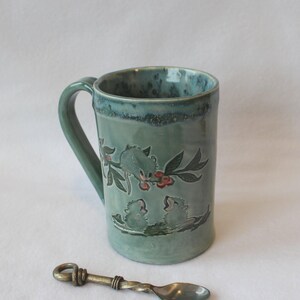 May include: A cylindrical ceramic mug with a glossy sage green glaze and a dark blue rim. The mug features a relief design of three small creatures on branches with red berries. A small, antique-style metal spoon with a twisted handle lies in front.