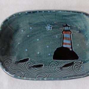 May include: An oval ceramic dish with a blue-green glaze featuring a lighthouse on a dark rock, a star, and three small whales swimming in embossed waves. The rim is decorated with a series of small, light blue dots around the entire edge.
