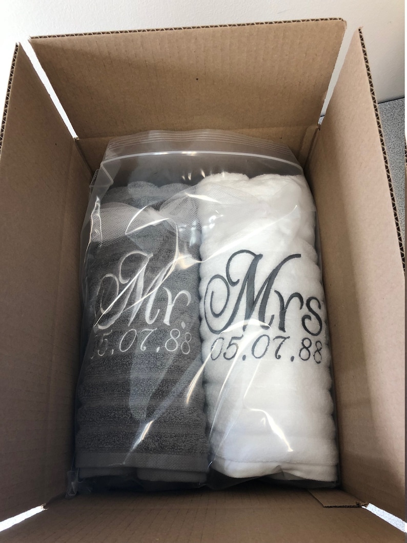 May include: Two rolled towels, one dark gray and one white, packaged in a clear plastic bag. The gray towel is embroidered with the text "Mr." and "05.07.88" in white, while the white towel features the text "Mrs." and "05.07.88" in dark gray.