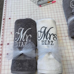 May include: Two rolled bath towels, one dark gray and one white, each embroidered with the text "Mr. DIAZ" and "Mrs. DIAZ" in a cursive script. Each towel is tied with a white tulle ribbon bow. A rotary cutter and a pink pen are visible nearby.