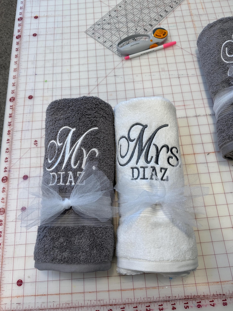 May include: Two rolled bath towels, one dark gray and one white, each embroidered with the text "Mr. DIAZ" and "Mrs. DIAZ" in a cursive script. Each towel is tied with a white tulle ribbon bow. A rotary cutter and a pink pen are visible nearby.