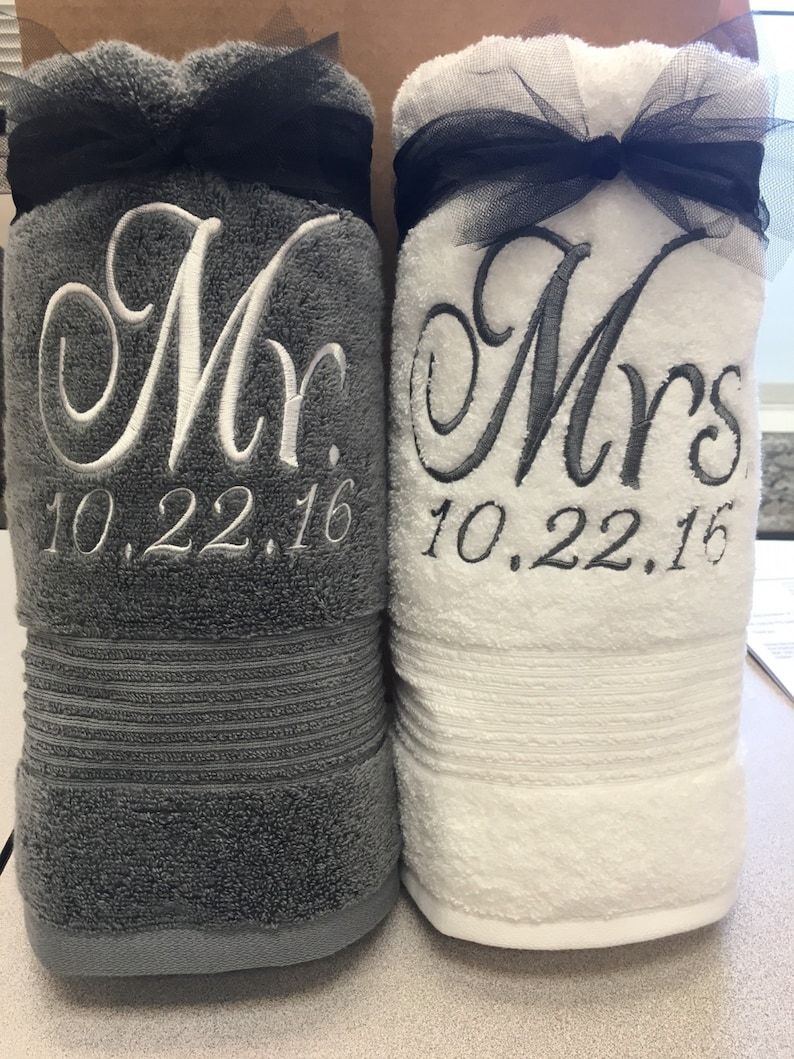 May include: Two rolled bath towels, one dark gray and one white, each tied with a black tulle ribbon. The gray towel is embroidered with white text reading Mr. and 10.22.16. The white towel is embroidered with dark gray text reading Mrs. and 10.22.16.