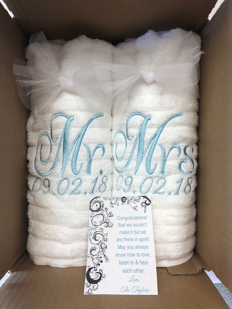 May include: Two white towels rolled and tied with white tulle bows, embroidered with light blue script reading Mr. and Mrs. above the date 09.02.18. A white card with black swirling patterns and text reads: Congratulations! Sad we couldn't make it but we are there in spirit! May you always know how to love, listen to & heal each other. Love, The Taylors.